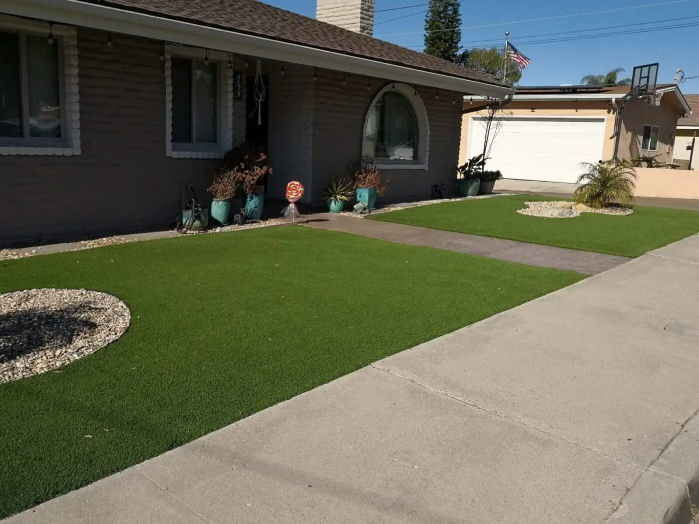 A neatly maintained front yard of a single-story house features artificial grass replacing the lawn, decorated with potted plants near the windows and entrance. A driveway with turf and pavers leads to a white garage door, and an American flag is visible in the background.