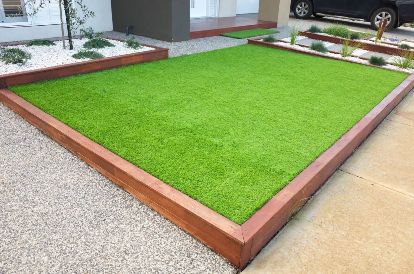 A well-maintained rectangular lawn, possibly featuring artificial turf, is bordered by reddish-brown wooden planks. The surrounding area includes gravel pathways and small garden beds with white stones and sparse green plants. A black car is partially visible in the top right corner.