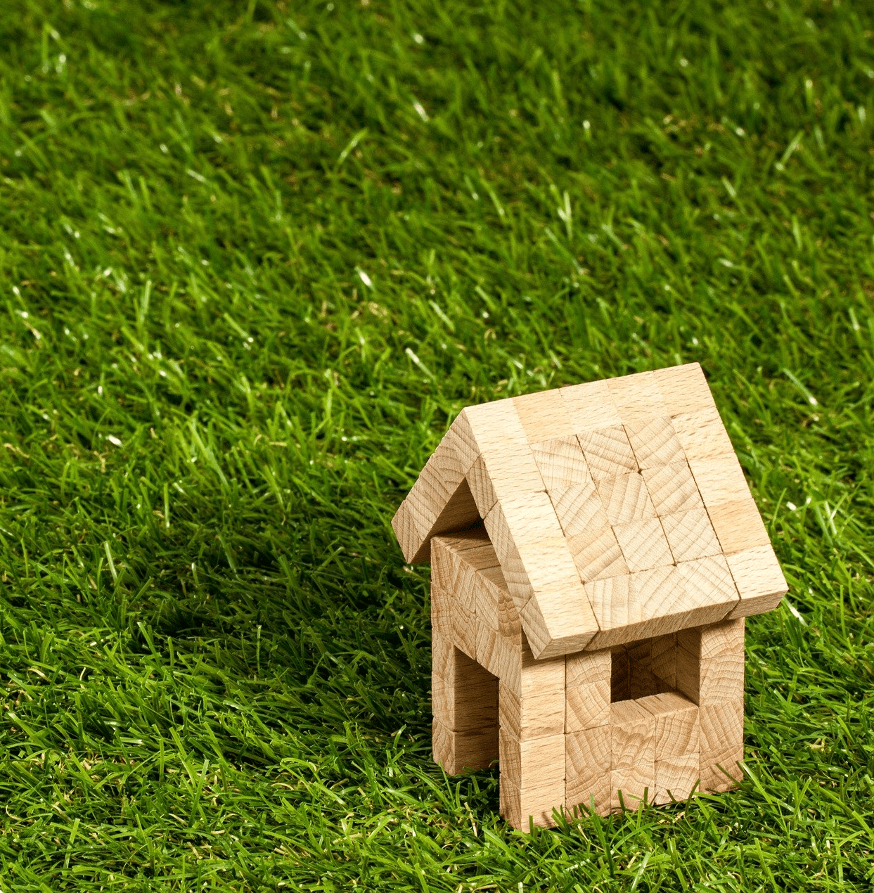 A small wooden toy house with a gabled roof stands on a patch of lush green grass. The house is made of light-colored wooden blocks, creating a simple, rustic structure. The background is filled with evenly cut pet-friendly artificial turf, giving a sense of an outdoor setting in Gilbert, AZ.