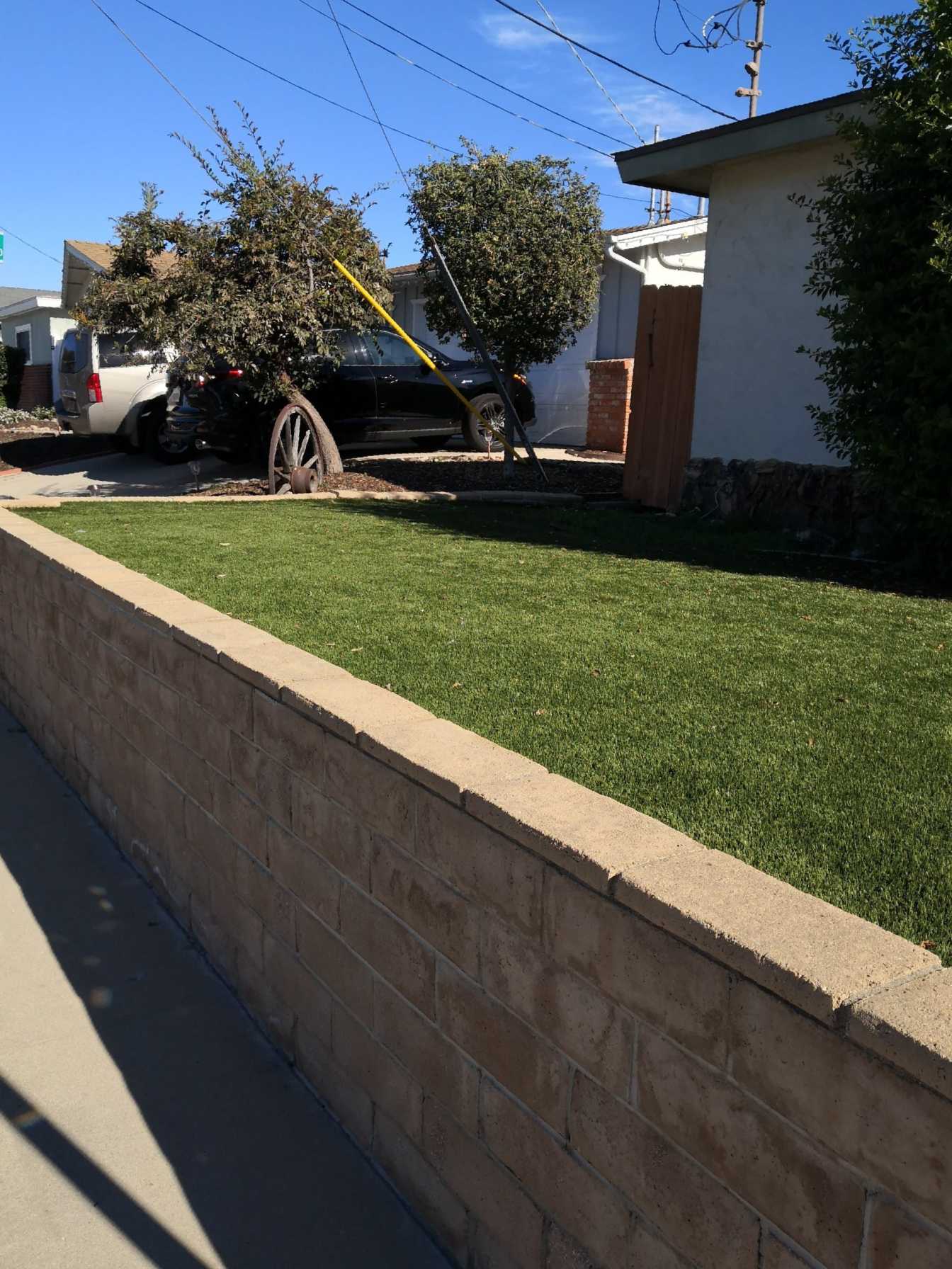 A suburban yard enclosed by a low brick wall, with lush, green pet turf. There is a black car parked in the driveway near a wagon wheel decoration, trees, and a light-colored house in the background. A clear blue sky is overhead. An artificial turf installer ensured the lawn stays vibrant year-round.